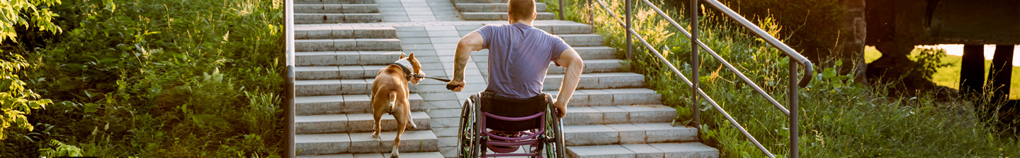 A man in a wheelchair with his dog, going up a ramp beside some stairs in a park
