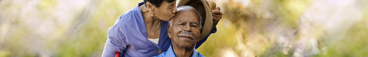 An elderly black gentleman on oxygen therapy being kissed on the head by his female caregiver