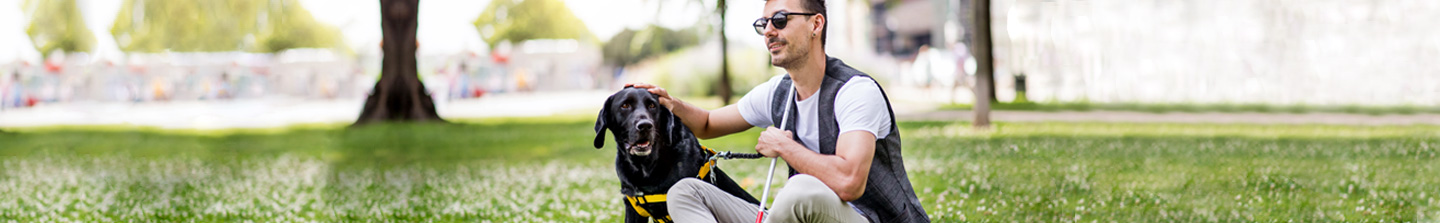 A 30-something blind man sitting on the grass in a park with his black lab guide dog at his side